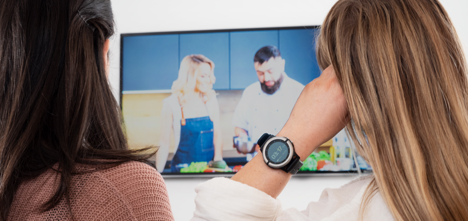 Image shows two people sitting on a couch enjoying a TV show about cooking. One woman is wear the Vitruvian Shield Monitoring watch on her wrist.