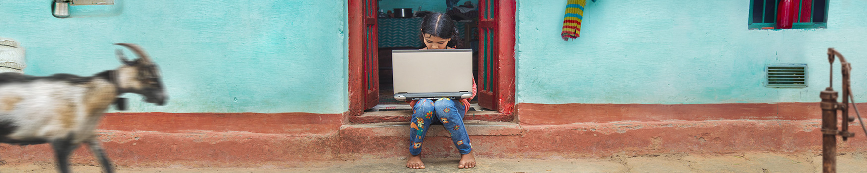 Child sitting on a step in a doorway holding an outdated laptop in a small village in India.