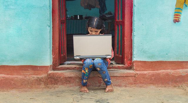 Child sitting on a step in a doorway holding an outdated laptop in a small village in India.