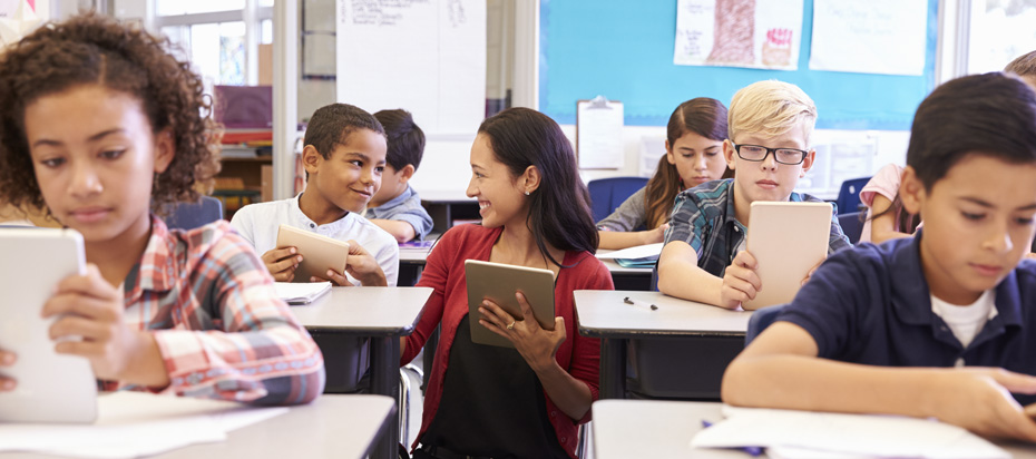 Middle school students at their desks holding mobile devices and a female teacher is kneeling next to one of the students to review their homework.