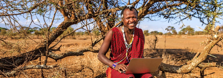 African tribe woman sits with a laptop computer on her lap. In the background is an African plains landscape.