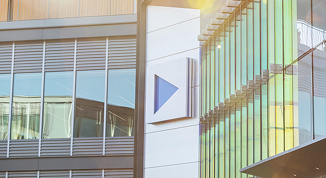 Modern corporate building with a blue triangular logo on the facade, featuring large glass windows reflecting sunlight and sleek architectural design.