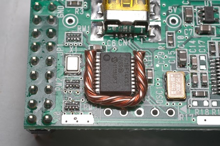 電子基板上のICチップ周辺に施された銅線の取り付け例。中央に黒いICチップがあり、その周囲に「コ」の字型に折り曲げられた茶色の銅線が巻かれています。電子工作やハンダ付けの解説に使用される写真で、SOP品の取り外し手順を示す技術的な参考画像です。