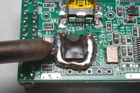 電子基板上でのハンダごてによるハンダの加熱作業の様子。ハンダごてが基板上のICチップ部分に当てられ、ハンダが溶けている状態を示しています。SOP部品の取り外し手順における重要なステップで、太く短いハンダごてを使用した効率的な加熱方法を示す技術的な画像です。